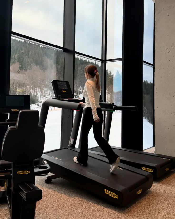 Woman exercising on a treadmill in gym with scenic snowy landscape view through large windows.