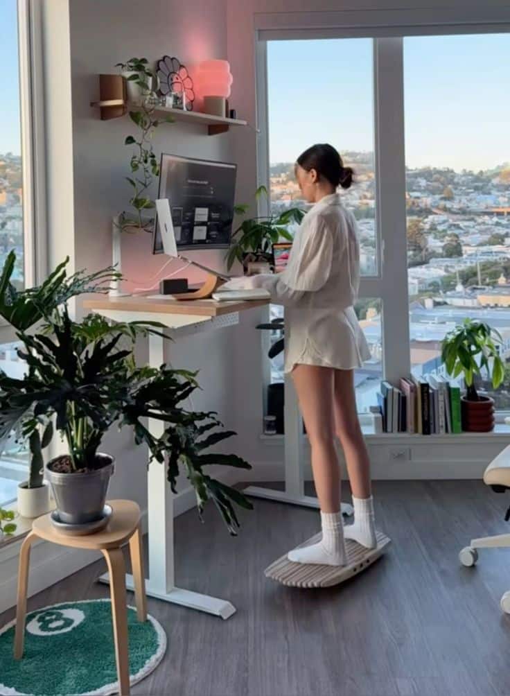 Woman working at a standing desk with a balance board, surrounded by plants, in a modern home office with city view.