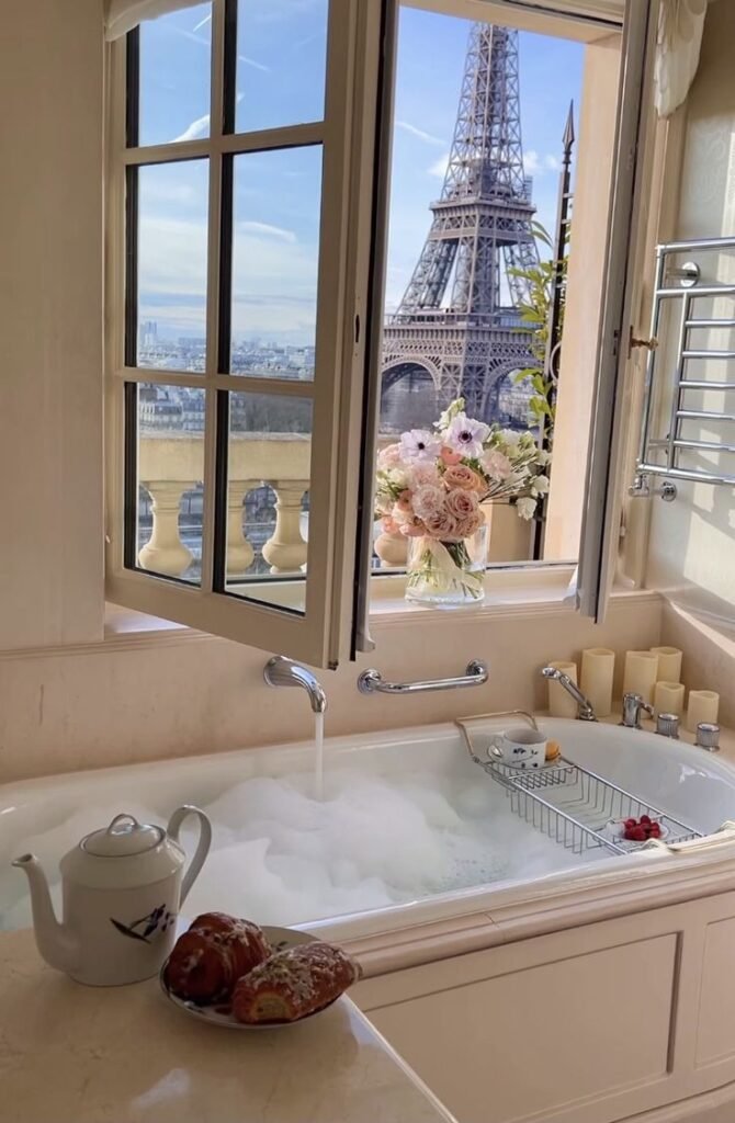 Luxurious Paris bathroom with Eiffel Tower view, featuring a bubble bath, flowers, and breakfast pastries by the window.