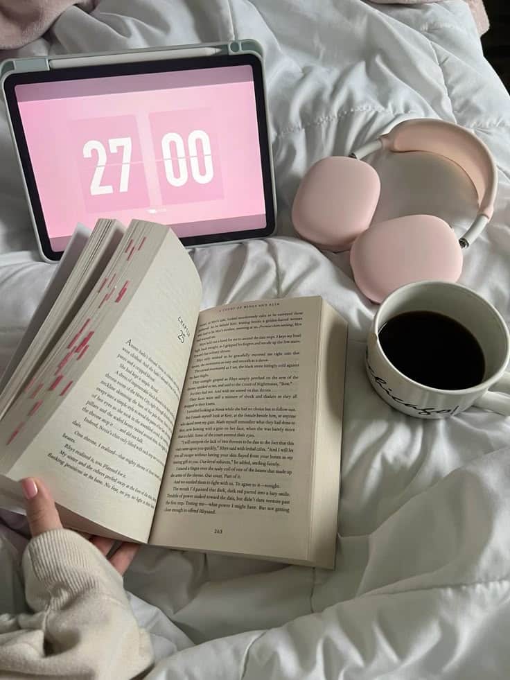 Cozy reading setup with an open book, pink headphones, timer on tablet, and a coffee mug on a white bedspread.