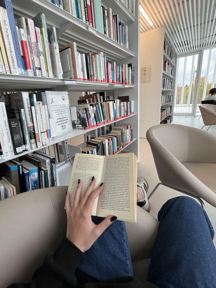 Person reading a book in a modern library with shelves of books and bright natural light coming through large windows.