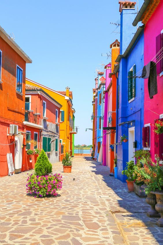 Venice landmark Burano island canal colorful houses italy - Handy Home Colorful houses on a charming cobblestone street in Burano, Italy, with potted plants lining the way under a clear blue sky.
