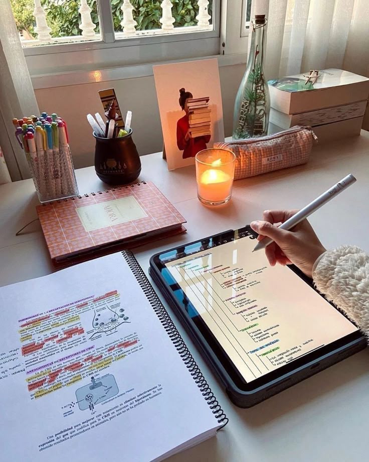 Study session: Tablet note-taking, colorful pens, and textbook on a cozy desk by the window with a warm candle ambiance.
