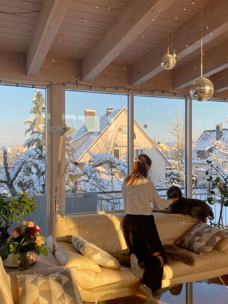 Cozy living room with a woman petting a dog by a window, snow-covered trees outside, warm interior decor, winter sunlight streaming in.