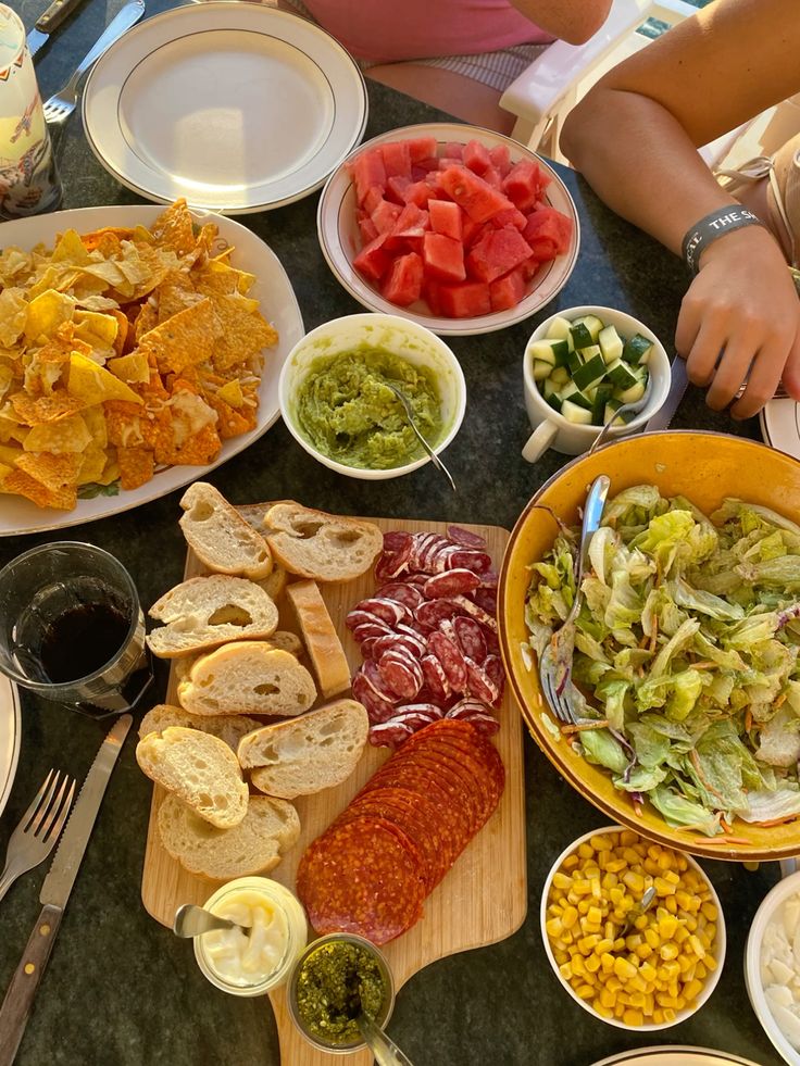tapas🇪🇸 - Handy Home Assorted snacks including chips, watermelon, guacamole, salad, corn, cured meats, and bread on a table, perfect for a summer gathering.