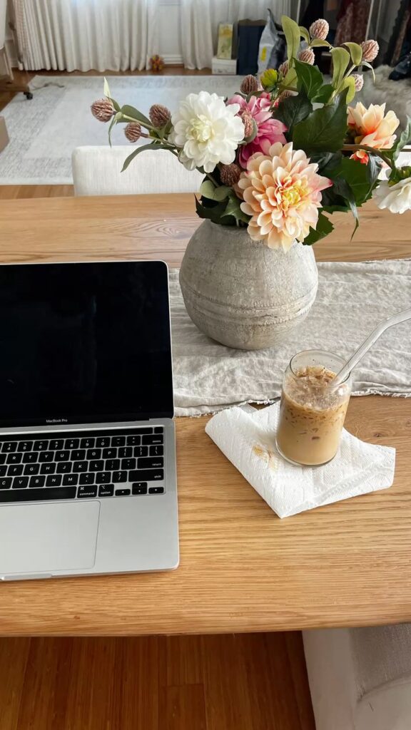Laptop on wooden desk with iced coffee and floral centerpiece, creating a cozy and productive workspace ambiance.