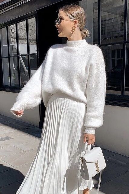 Woman in chic white sweater and pleated skirt, carrying a stylish handbag, showcases elegant street fashion.