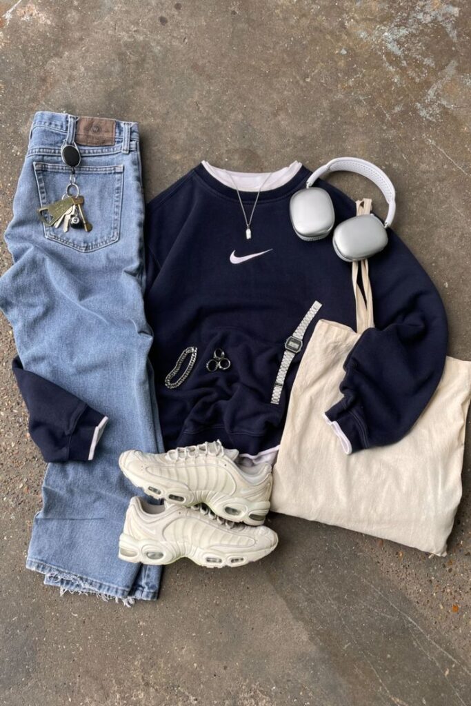 Casual outfit flat lay: navy sweatshirt, light blue jeans, white sneakers, headphones, tote bag, accessories on concrete floor.