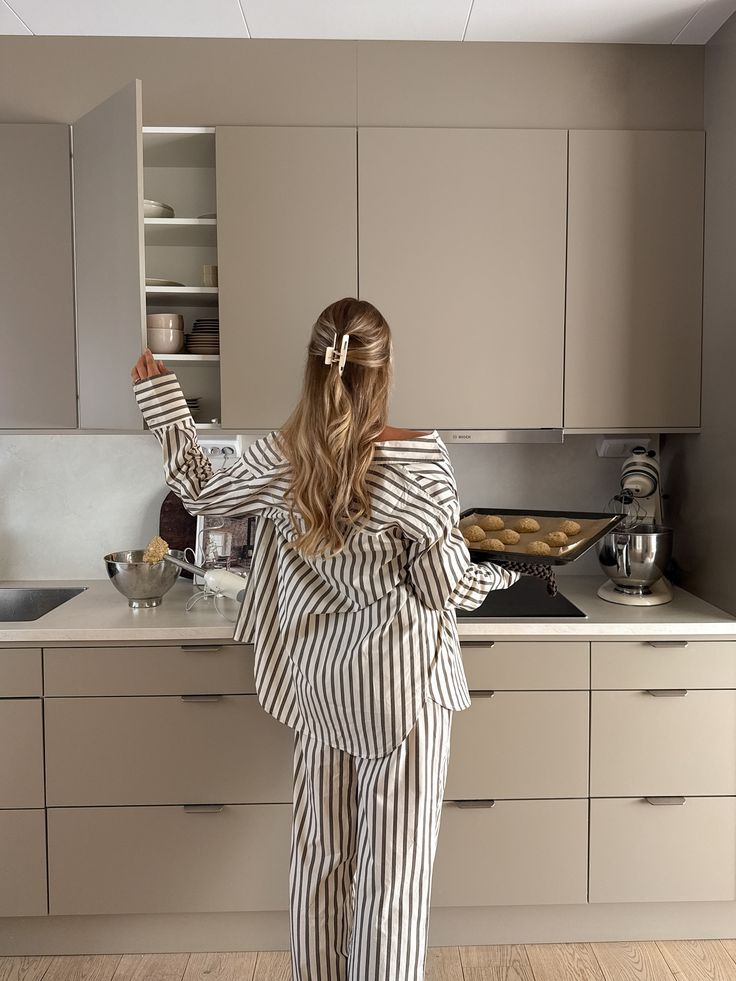Woman in striped pajamas holding a baking tray, reaching for open kitchen cabinet. Cozy home baking scene.