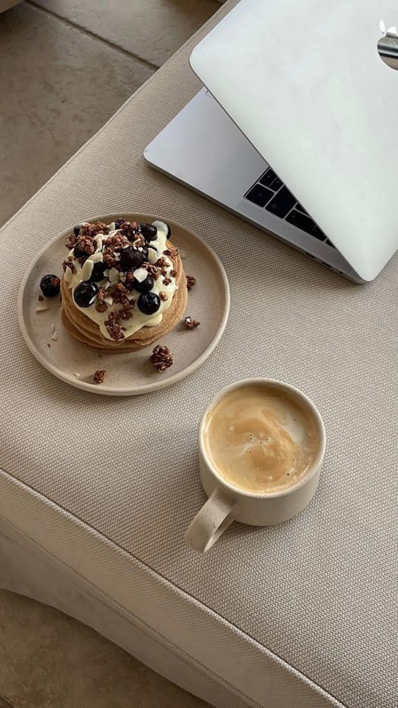 xfgnzdg - Handy Home A cozy setup with pancakes topped with cream and berries beside a frothy coffee and laptop on a beige fabric surface.