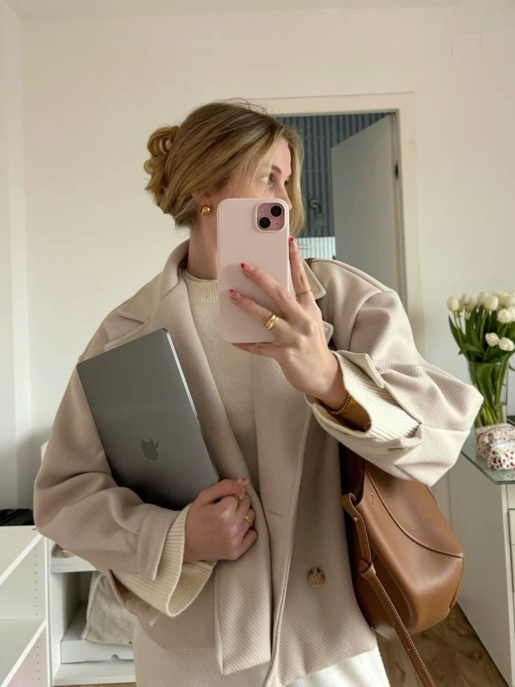 xfnxfn - Handy Home Woman in beige coat taking a mirror selfie with smartphone, holding a laptop, and carrying a brown bag, showcasing chic style.