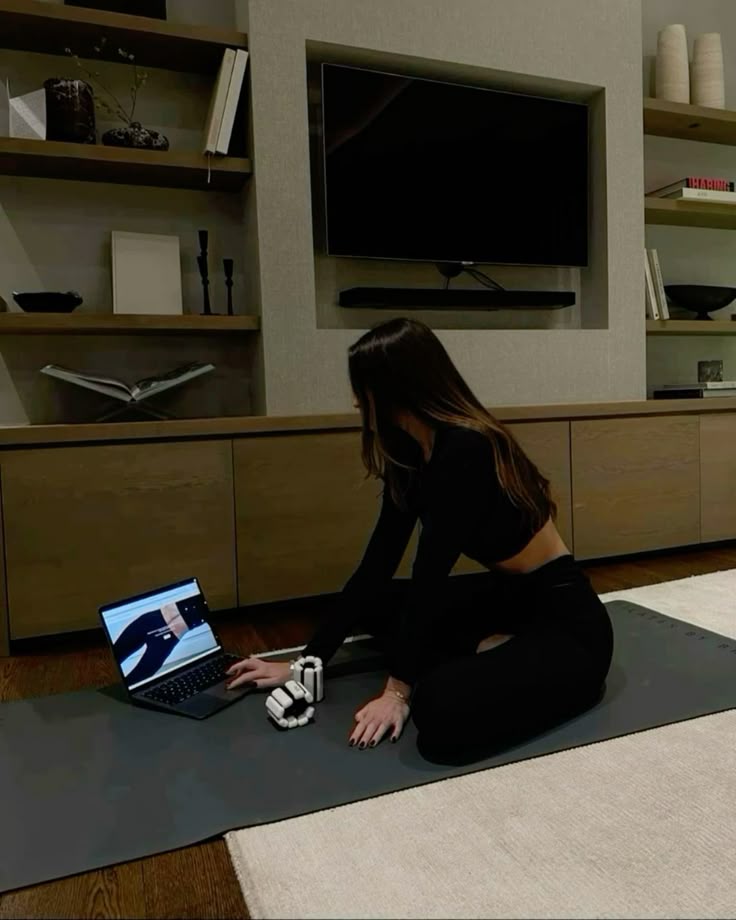Person practicing yoga at home on a mat, using a laptop for guidance, in a modern living room setup.