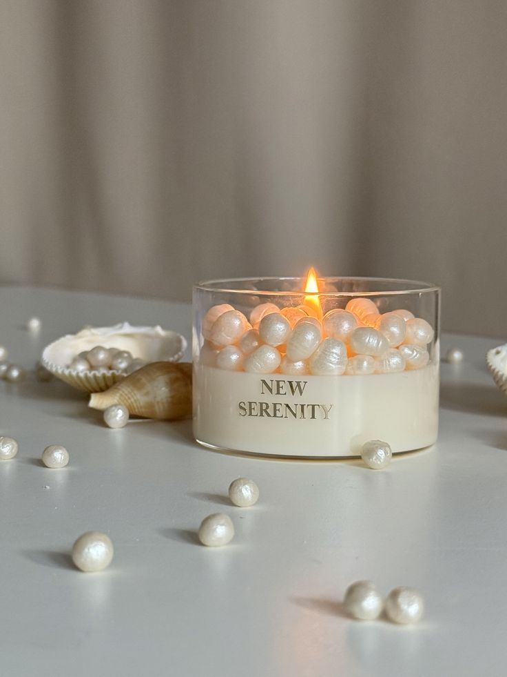 Lit candle labeled New Serenity decorated with pearls, surrounded by seashells, creating a calming ambiance.