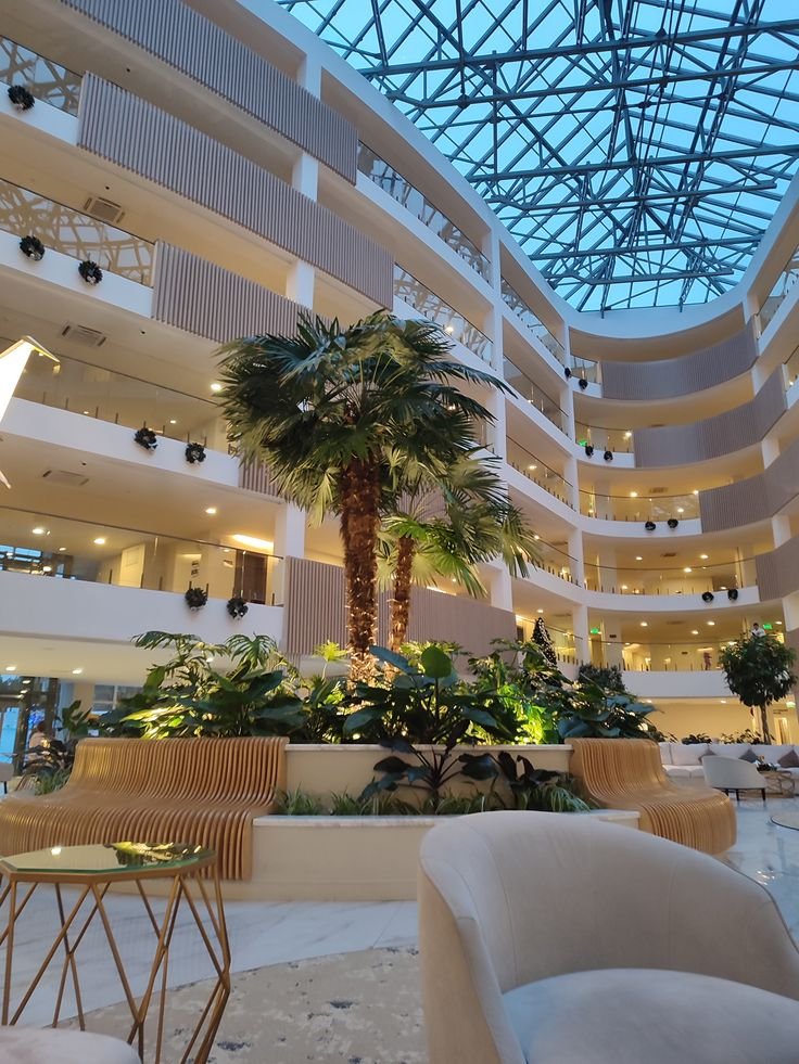 Luxurious hotel atrium with modern architecture, glass ceiling, indoor palm trees, elegant seating, and ambient lighting.