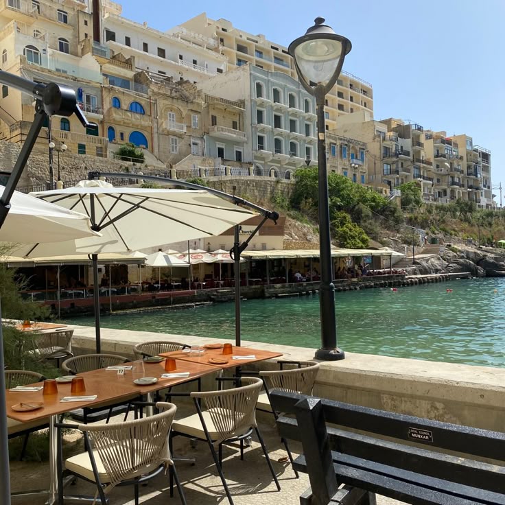 павияваи - Handy Home Riverside cafe with shaded tables by tranquil water, lined with charming European buildings under a clear blue sky.