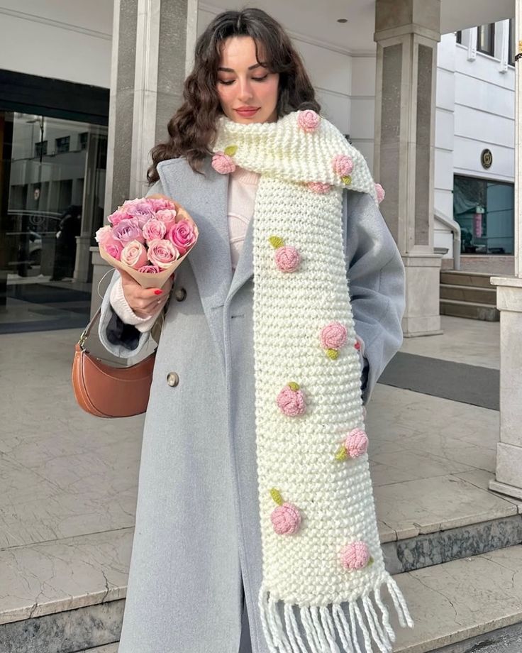 Woman in a light-blue coat with a knitted floral scarf holds a bouquet of pink roses.