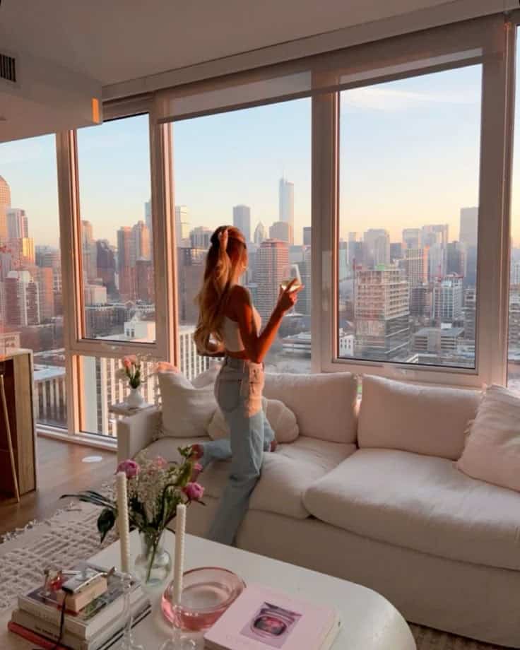 Woman enjoying sunset view of city skyline from a modern apartment living room. Cozy interior with books and flowers.