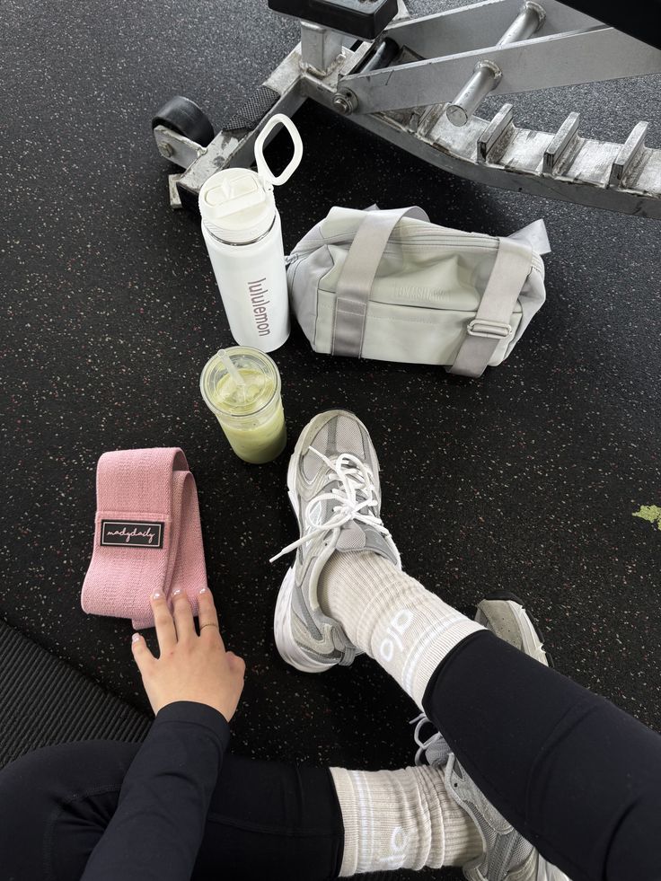 🍵 - Handy Home Person in gym sitting with workout gear, resistance band, sneakers, and drinks, ready for exercise session on black flooring.