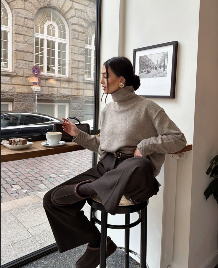 Screenshot - Handy Home Woman in cozy cafe, wearing a beige turtleneck and brown pants, enjoying coffee by a window with a city view. Elegant and relaxed vibe.