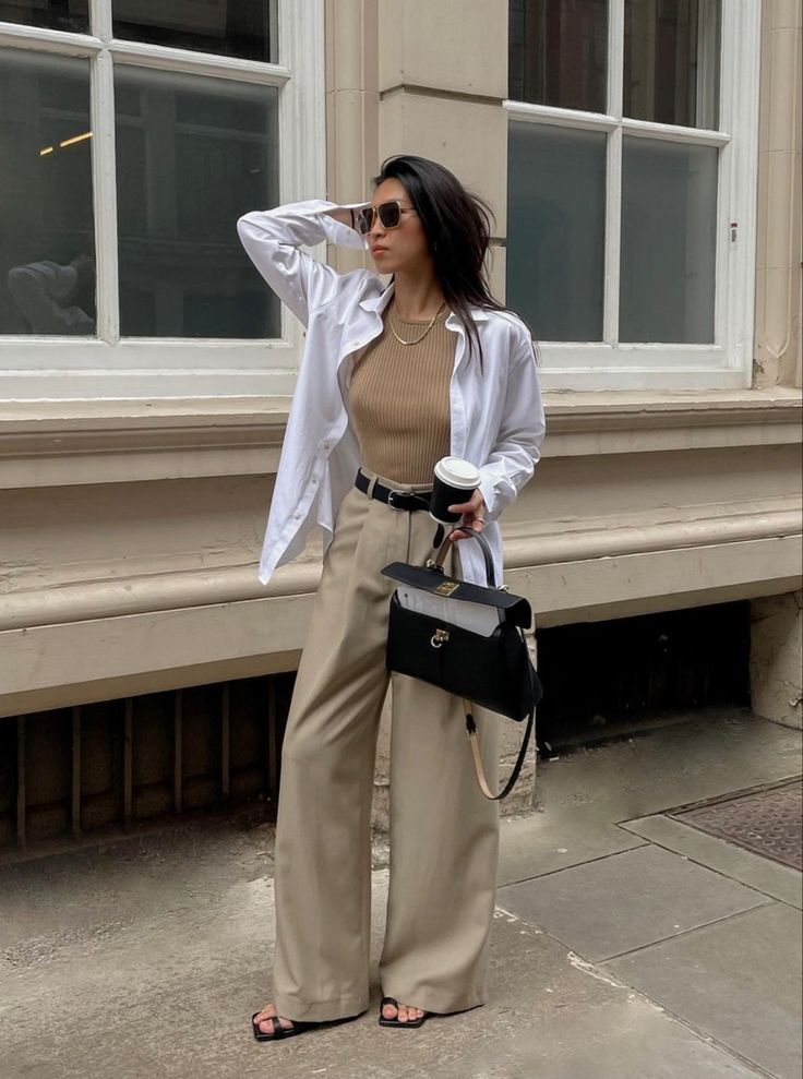 07903530-dd34-4fac-9fcb-6398d85983ce - Handy Home Stylish woman in beige trousers and white shirt, holding coffee and handbag, poses confidently on city street. Urban fashion vibe.