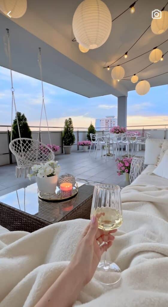 0ab5403d-9c58-45e0-9441-1fdac160be50 - Handy Home Cozy rooftop terrace with a blanket, a hand holding a wine glass, hanging lights, plants, and a sunset view. Perfect relaxation spot.