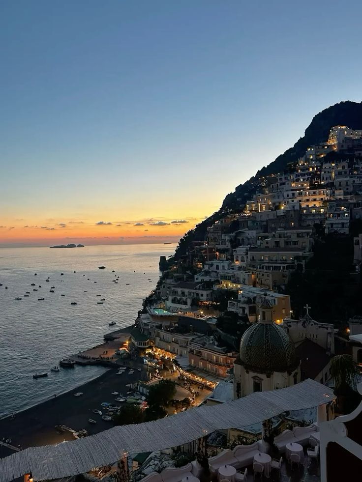 105b2468-b82b-46a9-8df3-26b097431a92 - Handy Home Scenic sunset view of Positano, Italy with illuminated hillside buildings and boats dotting the tranquil sea.