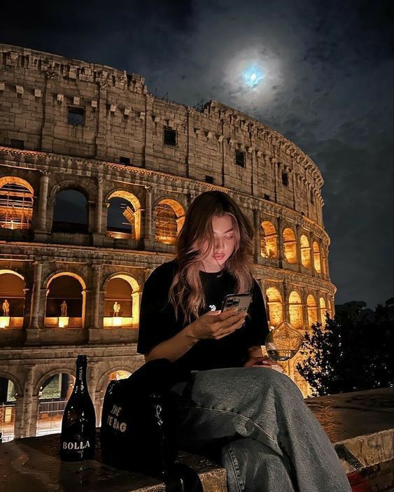1a408b0b-5eaa-4811-b9ec-13b727fdceee - Handy Home Woman using phone at night by illuminated Colosseum, full moon above, wine bottle on the side.