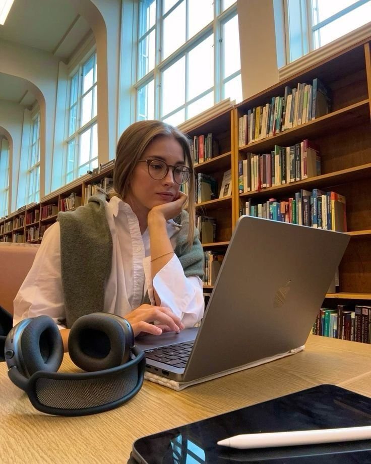 Person in a library using a laptop for study with headphones nearby, surrounded by bookshelves and large windows.