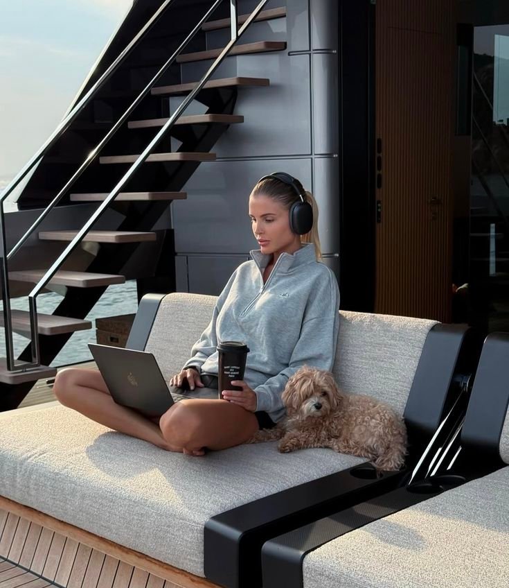 Woman working on a laptop with headphones, sitting on a yacht with a coffee and dog, enjoying a peaceful moment outdoors.
