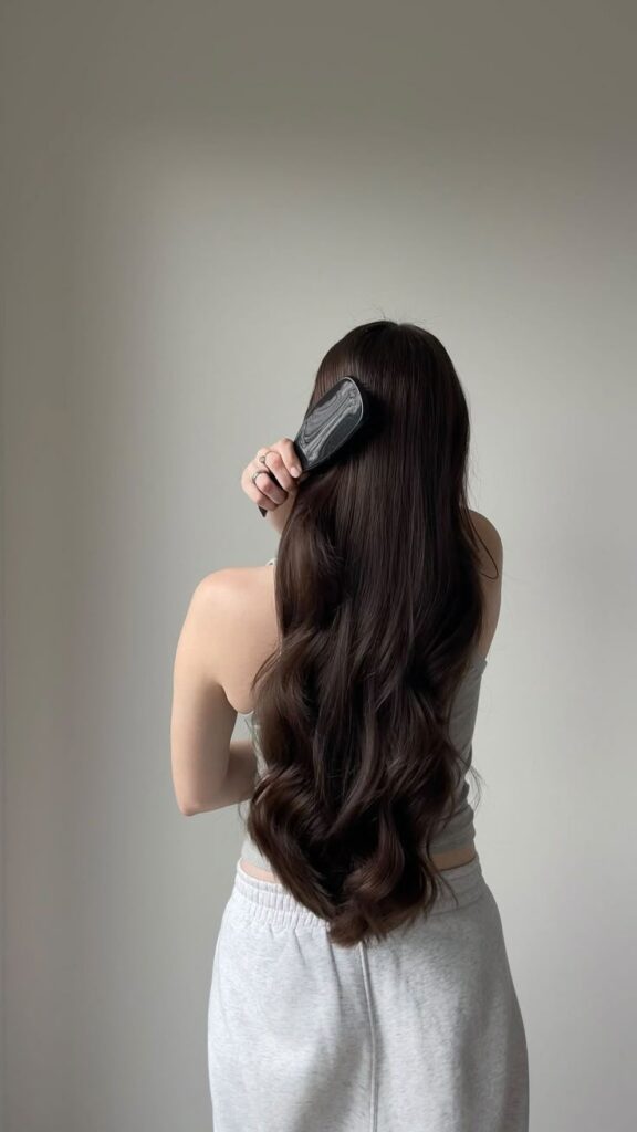 220ac89d-3380-4a2d-bd1a-a5e36812086d - Handy Home Woman brushing long, wavy brown hair in gray outfit. Hair care, daily routine, back view.