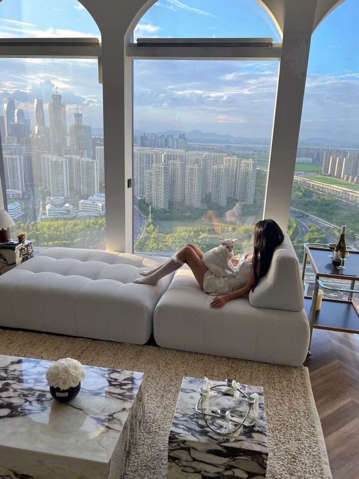 23-1765 Stylish Home Design Ideas for Every Room - Handy Home Woman and cat relaxing on a modern sofa with a stunning cityscape view through large arched windows. Luxurious urban apartment.