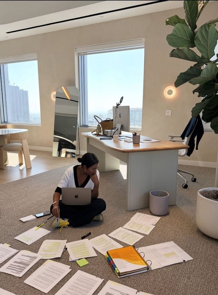 Person working on a laptop, surrounded by documents on the floor in a modern office.