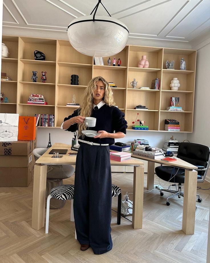 35ba9b90-aefe-4afd-a746-0e8385c8823c - Handy Home Stylish woman in a modern office holding coffee, surrounded by books and decor on shelves, under a large ceiling light.