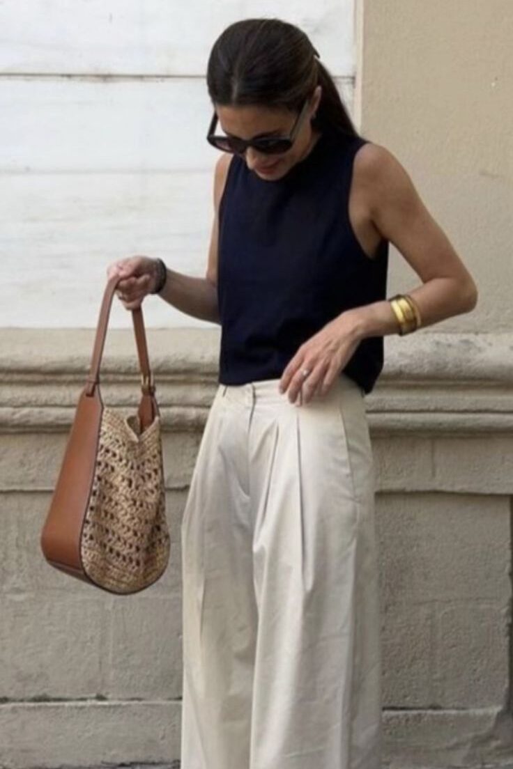 3e8d8f12-9cc2-4f53-8fc7-e8280ca83b67 - Handy Home Woman in stylish black top, beige wide-leg pants, holding a woven handbag. Chic summer fashion outfit.