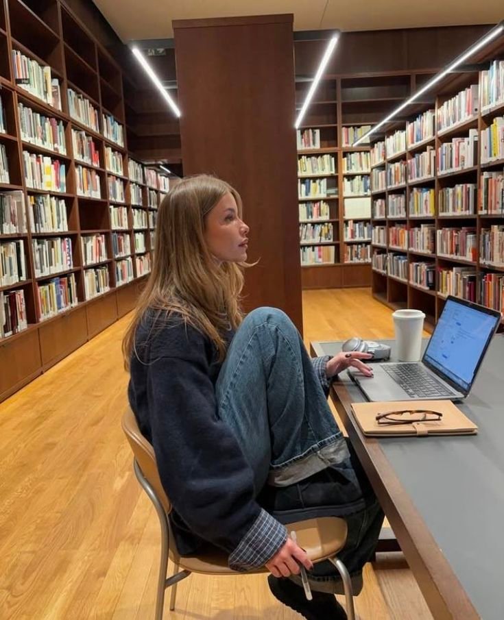 Young woman studying in a library with a laptop and notebook, surrounded by shelves of books. Stylish casual outfit.