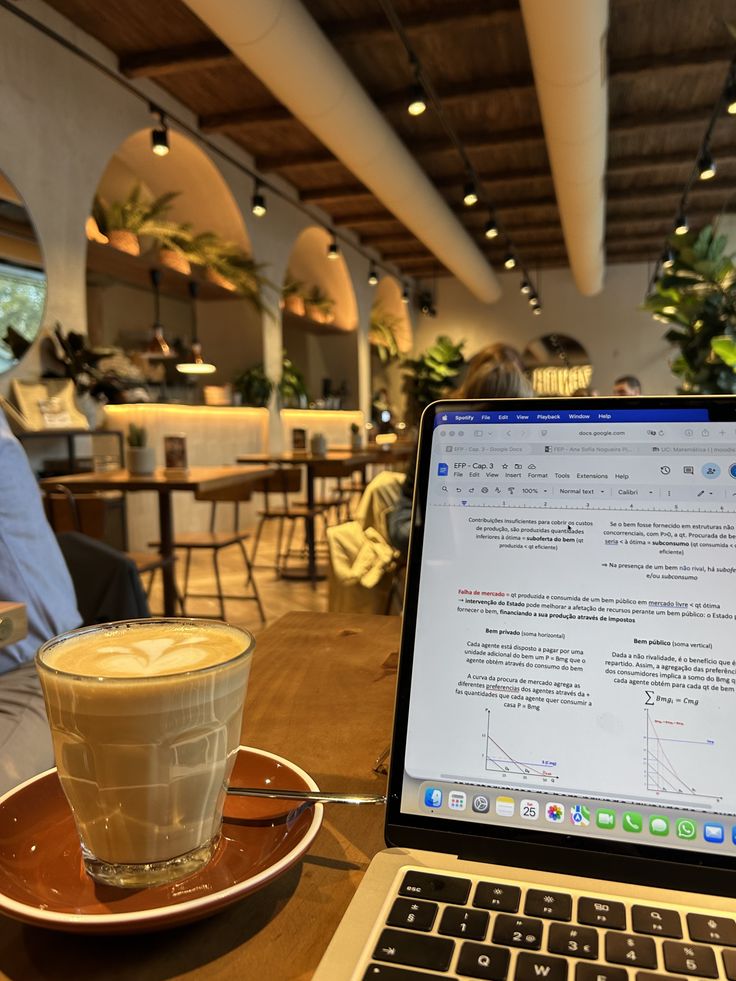 4735c166-4032-4e78-abc6-86b620d9347a - Handy Home Laptop with open document and latte on a café table, surrounded by cozy seating and greenery. Perfect setting for work or study.