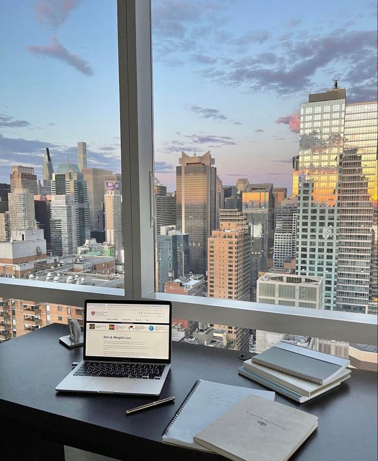 5ae24738-1d9c-48d8-9e82-419e36407619 - Handy Home Laptop on desk with notebooks, pen, overlooking a stunning city skyline at sunset through a large window.