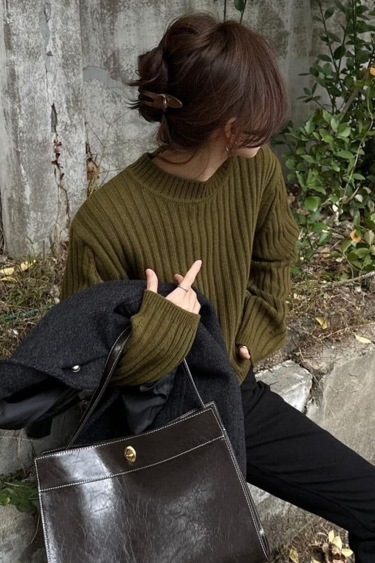 62b2facf-895f-445e-a197-495af80889fa - Handy Home Fashionable woman with brown leather handbag and olive sweater, sitting outdoors against a stone wall, surrounded by greenery.