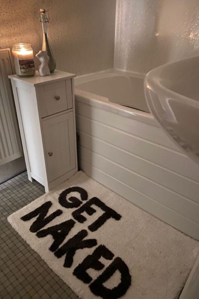 Modern bathroom decor with a Get Naked bath mat, candle, and small cabinet. Cozy and humorous touch to the bathing area.