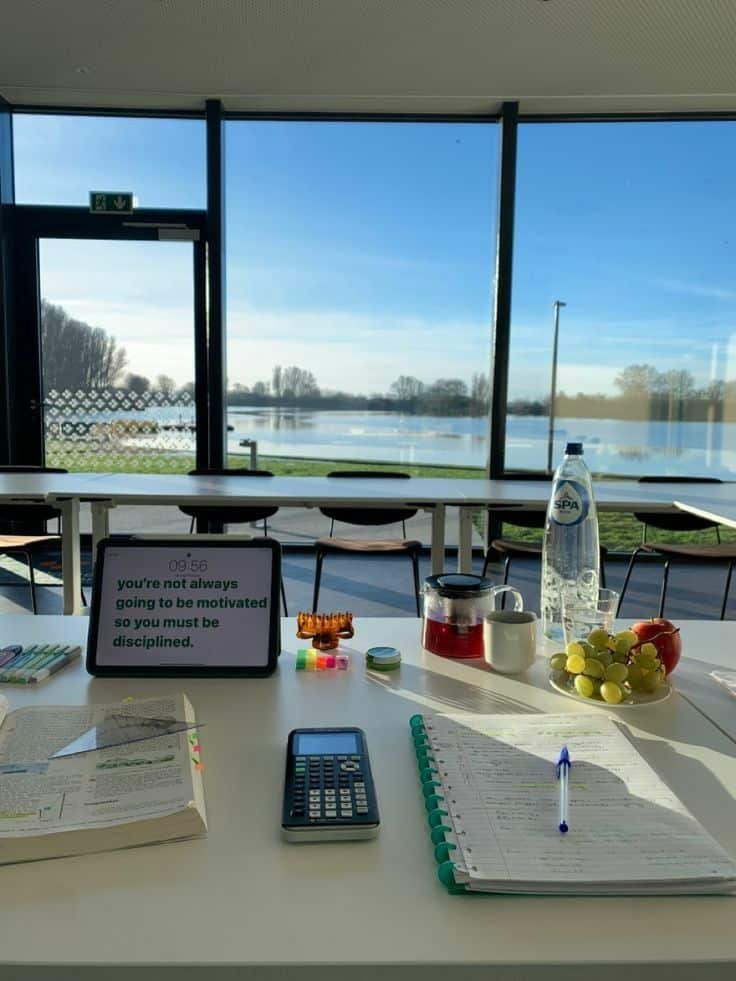 Study setup by a bright window with notes, calculator, fruit, tea, and motivational quote on tablet.