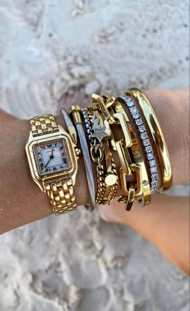 7d065bb4-54b4-4060-88d0-93dbf2628614 - Handy Home Stylish wrist adorned with gold watch and bracelets against a sandy beach backdrop, showcasing elegant jewelry fashion.