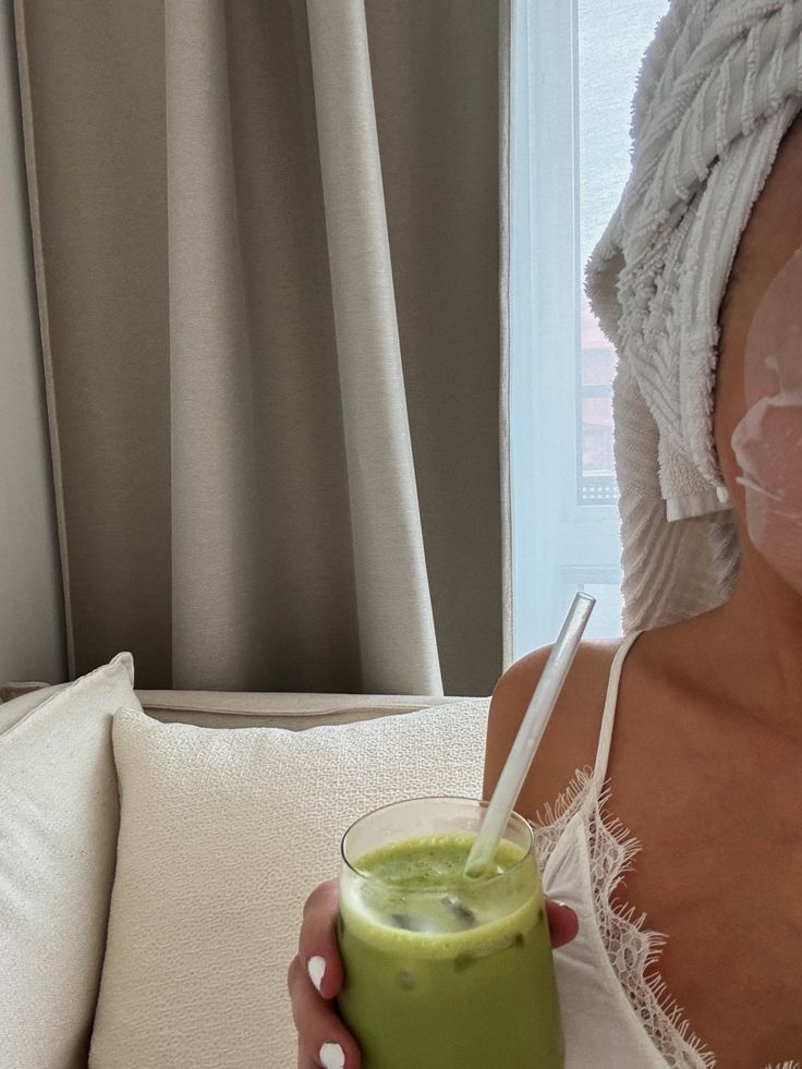 7e8bb2ee-04cc-49ed-b764-62f5582cb66b - Handy Home Woman enjoying iced green drink, wearing towel and eye patches, relaxing on sofa in cozy room.