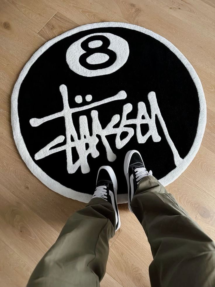 Person standing on round rug with a bold design and number eight, wearing khaki pants and black sneakers. Modern streetwear style.