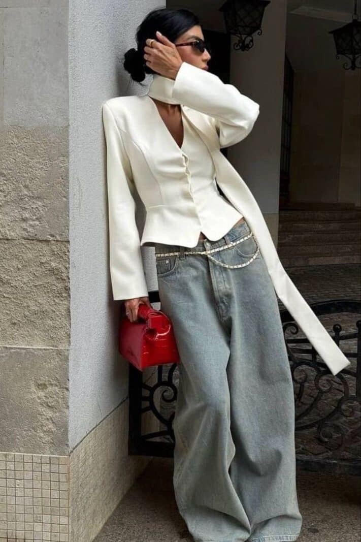 82079680-de87-4a5b-b484-c59b46894f93 - Handy Home Stylish woman in white blazer and wide jeans holding red bag, leaning against wall, exuding chic urban fashion vibes.