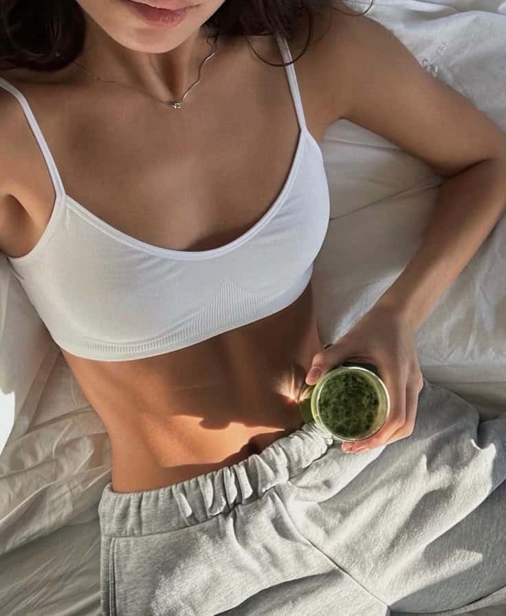 84686040-0726-461a-9477-35ef3217b62f - Handy Home Woman in white top and grey pants holding a green smoothie while lounging on bed, promoting healthy lifestyle and relaxation.