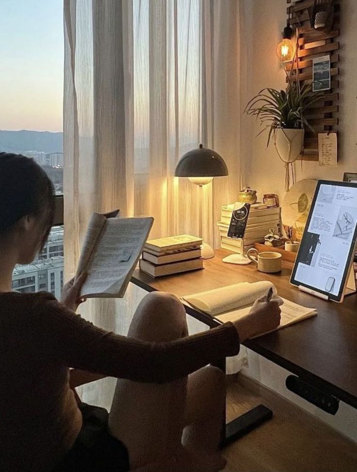 84849937-ce33-4343-a87a-d58d896d6aab - Handy Home Cozy home office with books, tablet, and ambient lighting; woman reading by the window with a city view at sunset.