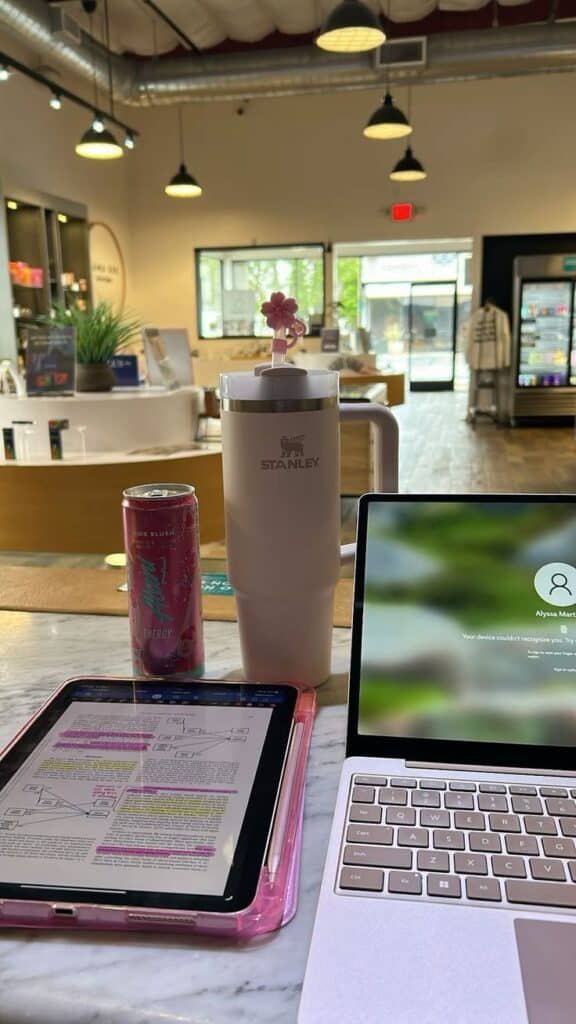 Coffee shop workspace with a laptop, tablet, energy drink, and a Stanley tumbler on a marble table, cozy and productive ambiance.