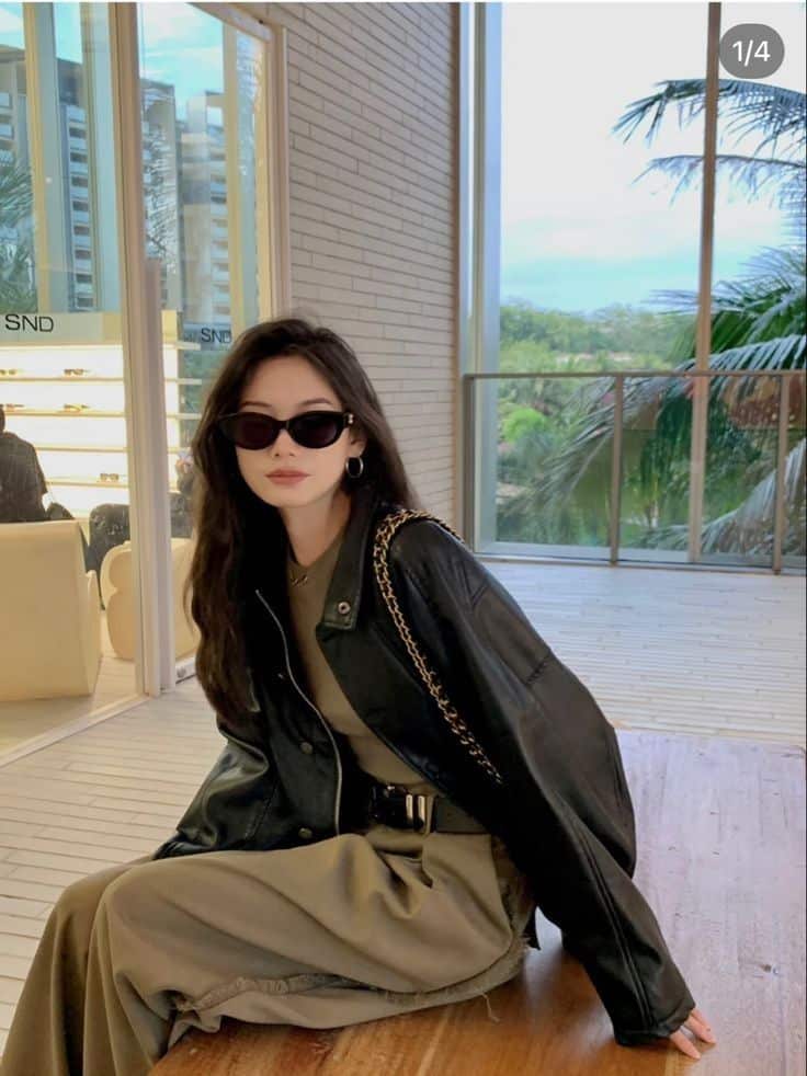 92126672-347a-4944-ba97-1e374ba42aa1 - Handy Home Stylish woman in sunglasses and black leather jacket, sitting indoors with palm trees visible through large windows.