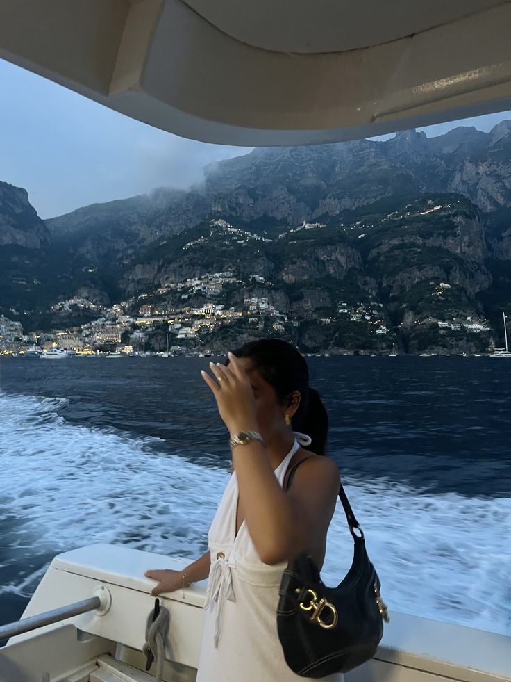 98af34ba-1968-4acc-8363-b2c8deeca943 - Handy Home Woman on boat with coastal view of a seaside town and towering mountains in the background at dusk.