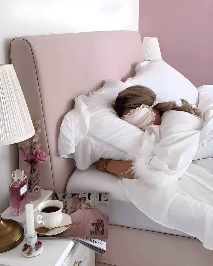 Cozy bedroom with pink decor, a woman sleeping in white linens, Vogue magazine, coffee, and flowers on a bedside table.